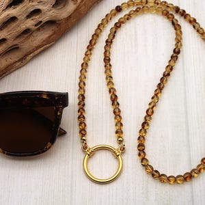 May include: A necklace with amber-colored beads and a gold-colored circular pendant. Tortoiseshell sunglasses and a piece of driftwood are also in the image. The logo "Crafted by Kristal" is in the lower left corner.