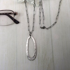 Puede incluir: Un collar plateado con un colgante ovalado con textura martillada. El collar se muestra con un par de gafas y un elemento decorativo de cristal. La imagen también incluye un logotipo que dice "Crafted by Kristal".