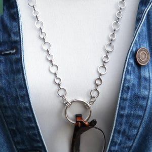 May include: A silver-toned eyeglass holder necklace with a series of connected circles. The necklace is holding a pair of tortoiseshell eyeglasses. The background includes a white shirt and a denim jacket with a copper-colored button.