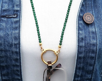 Könnte beinhalten: Eine grüne Perlenkette für eine Brillenhalterung mit einem goldfarbenen Ring und Verschluss. Die Kette wird über einem weißen Hemd und einer Jeansjacke getragen. Der Text "Crafted by Kristal" ist oben links sichtbar.
