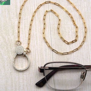 May include: A gold-colored eyeglass chain with a decorative clasp encrusted with small, clear stones. The chain is connected to a pair of eyeglasses with a brown frame. The image also includes a logo that says "Crafted by Kristal".