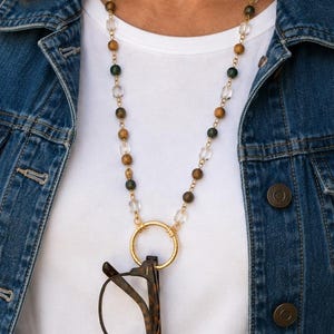 May include: A beaded eyeglass holder necklace featuring alternating brown tiger eye, dark green, and clear faceted beads on a gold-tone chain. The necklace includes a circular gold-tone ring pendant designed to hold a pair of tortoise shell glasses.