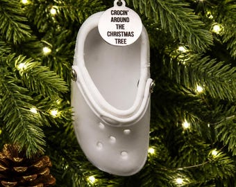 May include: A white foam clog ornament hanging from a silver string. A circular silver charm is attached to the top, featuring the black text 'CROCIN' AROUND THE CHRISTMAS TREE'. The clog has a smooth surface with ventilation holes and a heel strap.
