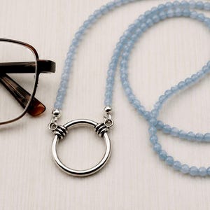 Op de afbeelding: Een lichtblauwe glazenketting met een zilveren ringhanger. De ketting wordt getoond naast een bril met bruin montuur. De afbeelding bevat ook een logo met de tekst "Crafted by Krystal".