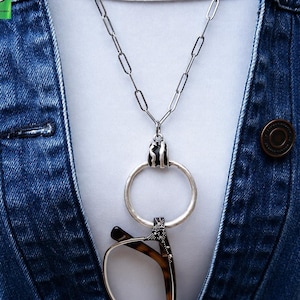 May include: A silver necklace with a large ring pendant holding a pair of spectacles. The necklace is worn over a white top and a denim jacket. The spectacles have a tortoiseshell pattern. The pendant has a black and white zebra stripe design.
