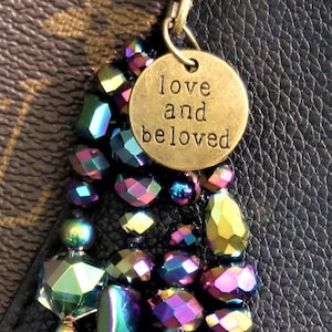 May include: A colorful iridescent beaded keychain with a gold charm that reads "love and beloved". The keychain is hanging from a brown leather bag.