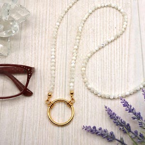 May include: A long necklace featuring small, iridescent white beads strung together, ending in a polished gold-tone circular ring pendant. The design includes a secure metal clasp mechanism connecting the beaded strand to the central gold ring.