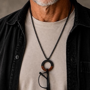 May include: A black chain necklace featuring a circular pendant made of two-toned wood, with a dark brown lower half and a black upper half. The pendant serves as a holder for a pair of black-rimmed spectacles, which hang securely from the ring.