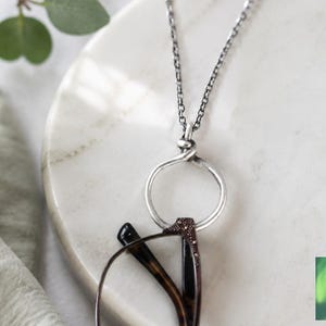 May include: A silver necklace with a circular pendant holding a pair of spectacles. The glasses have a brown and black frame. The necklace is on a white marble surface, with green leaves in the background. The image includes a logo that says "Crafted by Kristal".