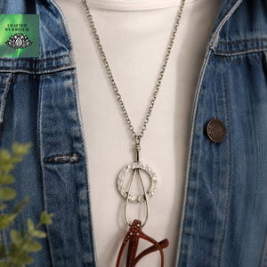 May include: A silver-toned necklace with a textured, circular pendant and a teardrop-shaped holder for eyeglasses. The necklace is worn over a white shirt and a denim jacket. The eyeglasses are brown.