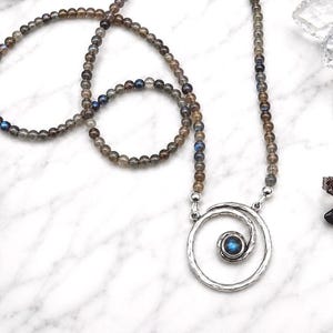 May include: A beaded necklace with a silver pendant featuring a spiral design and a blue gemstone center. The necklace is made of small, round, gray and brown beads. The image also includes a pair of eyeglasses and a green and white logo.
