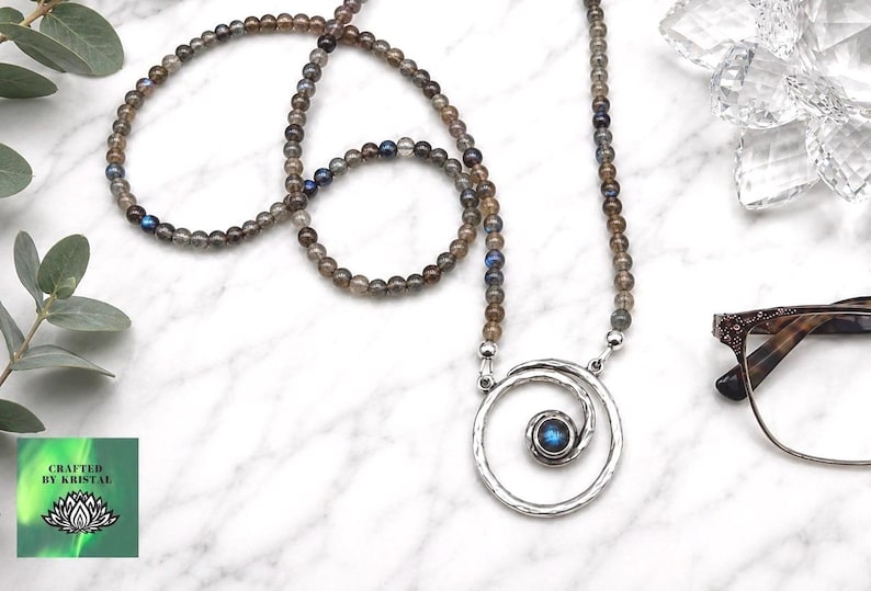 May include: A beaded necklace with a silver pendant featuring a spiral design and a blue gemstone center. The necklace is made of small, round, gray and brown beads. The image also includes a pair of eyeglasses and a green and white logo.