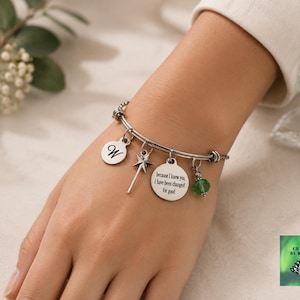 May include: Silver-toned charm bracelet with multiple charms. Charms include a letter "W", a fairy wand, a circular charm with the text "because I knew you, I have been changed for good", and a green bead. The bracelet is on a wrist.
