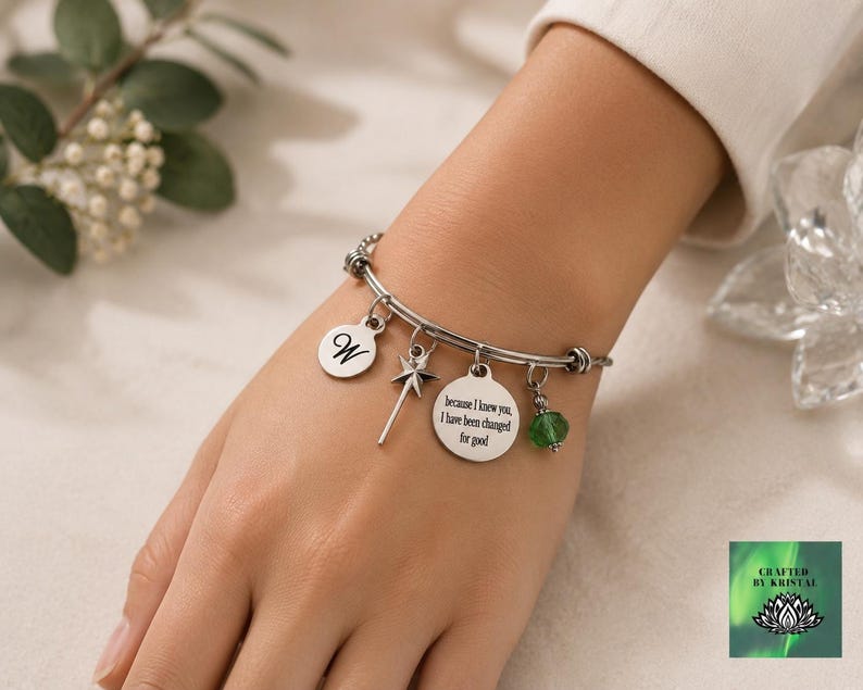 May include: Silver-toned charm bracelet with multiple charms. Charms include a letter "W", a fairy wand, a circular charm with the text "because I knew you, I have been changed for good", and a green bead. The bracelet is on a wrist.