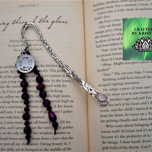 May include: Silver bookmark with a charm that says "Momlife" and a glass of wine with a wine bottle. The charm is attached to a strand of dark purple beads. The bookmark is laying on a book with the text "seeing through the glass" and "Chapter 7".