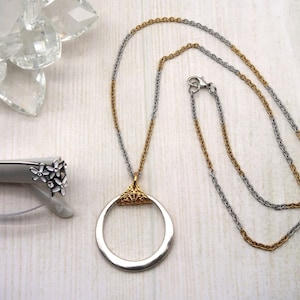May include: A silver and gold-toned necklace with a circular pendant. The chain is two-toned, with silver and gold links. The pendant has a silver ring with a gold filigree design at the top. The image also shows eyeglasses with floral accents.