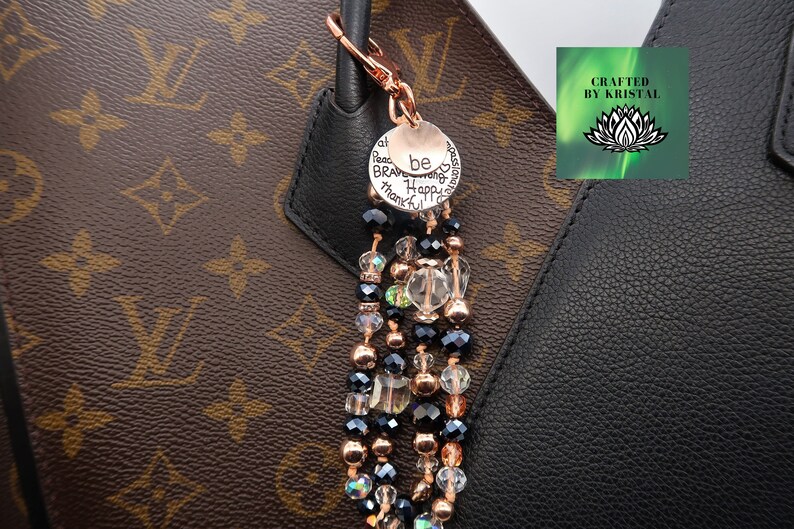 K&ouml;nnte beinhalten: Ein Gl&uuml;cksarmband mit einem ros&eacute;goldenen Anh&auml;nger, auf dem "be brave, be strong, be happy, be thankful" steht. Der Anh&auml;nger ist an einer Kette aus schwarzen und klaren Kristallperlen befestigt. Das Armband h&auml;ngt an einer braunen und schwarzen Handtasche.