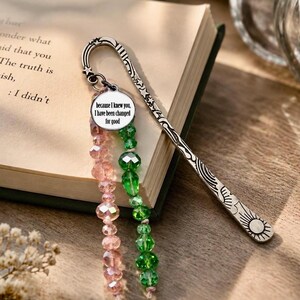 May include: A silver-toned metal bookmark featuring a celestial design with stars and a sun, attached to a circular charm with the text 'because I knew you, I have been changed for good' and two strands of faceted pink and green glass beads.