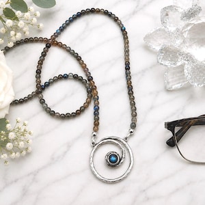 May include: A beaded necklace with a silver spiral pendant and a blue gemstone center. The necklace is displayed with a white rose, eyeglasses, and a decorative crystal flower on a marble surface.