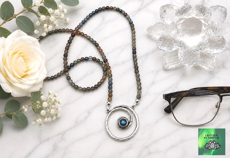 May include: A beaded necklace with a silver spiral pendant and a blue gemstone center. The necklace is displayed with a white rose, eyeglasses, and a decorative crystal flower on a marble surface.