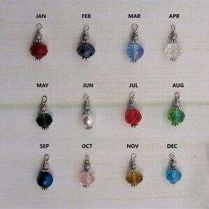 May include: A collection of twelve beaded charms, each representing a month of the year. Each charm features a faceted bead in a different color, with silver-toned metal accents and a loop for attachment. The months are labeled above each charm.