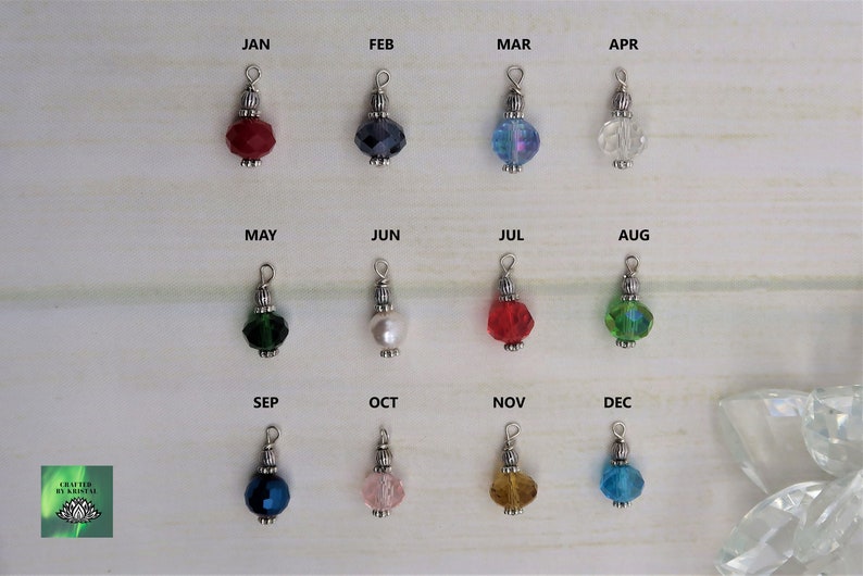 May include: A collection of twelve beaded charms, each representing a month of the year. Each charm features a faceted bead in a different color, with silver-toned metal accents and a loop for attachment. The months are labeled above each charm.