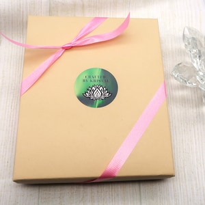Könnte beinhalten: Eine braune Geschenkbox mit einem rosa Band darum gebunden. Die Box hat einen Aufkleber mit einem grünen und schwarzen Lotusblumen-Design und dem Text "Crafted by Kristal".