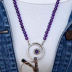 Peut inclure: Un collier composé d'un rang de perles rondes en améthyste violette relié à un pendentif en métal argenté en forme de spirale. Le centre de la spirale contient une petite pierre d'améthyste ronde. Le pendentif sert de support à des lunettes.