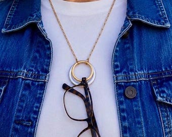 May include: A gold chain necklace with a pendant featuring two interlocking rings, one gold and one silver. Tortoiseshell eyeglasses are hanging from the pendant. The necklace is worn over a white shirt and a blue denim jacket.