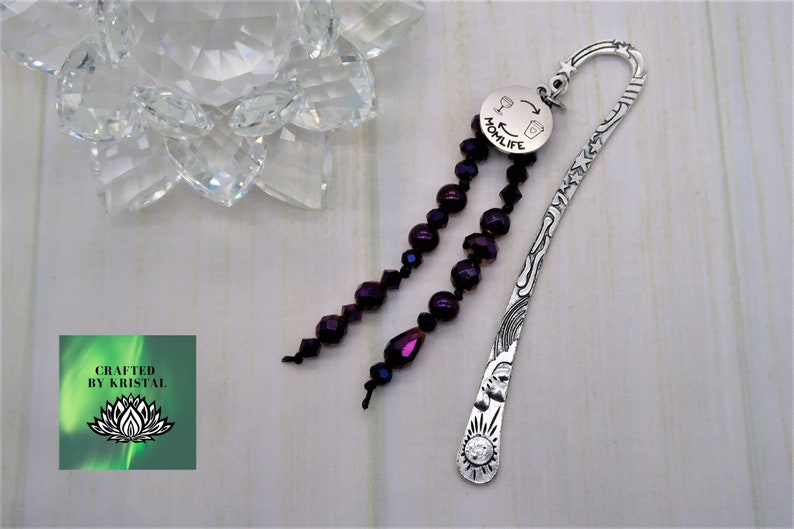 May include: Silver bookmark with a purple beaded tassel. The bookmark is engraved with a sun, stars, and a rainbow. The tassel is attached to a round charm that says "Momlife" with a wine glass and coffee cup graphic.