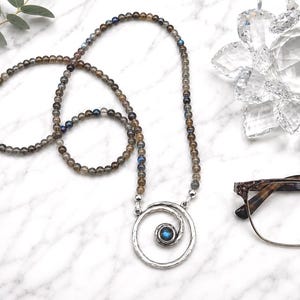 May include: A beaded necklace with a silver pendant featuring a spiral design and a blue gemstone. The necklace is made of small, round, gray and brown beads. The image also includes eyeglasses and a decorative crystal flower.