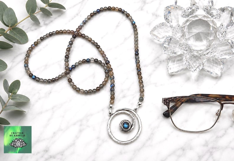 May include: A beaded necklace with a silver pendant featuring a spiral design and a blue gemstone. The necklace is made of small, round, gray and brown beads. The image also includes eyeglasses and a decorative crystal flower.