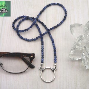 May include: A blue beaded necklace with a silver ring pendant. The necklace is made of small, round, blue beads. The pendant is a large, silver ring. The necklace is displayed with a pair of spectacles.