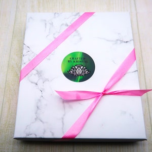 K&ouml;nnte beinhalten: Eine wei&szlig;e Box mit einem rosa Band, das darum gebunden ist. Die Box hat ein Marmormuster und einen Aufkleber mit der Aufschrift "Crafted by Kristal" mit einem gr&uuml;nen und schwarzen Lotusbl&uuml;ten-Design.