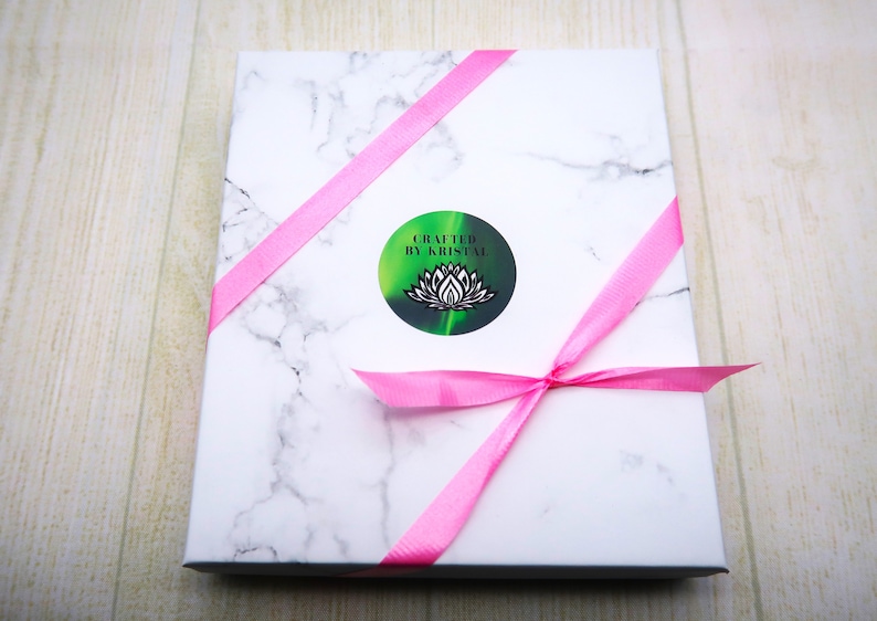 K&ouml;nnte beinhalten: Eine wei&szlig;e Box mit einem rosa Band, das darum gebunden ist. Die Box hat ein Marmormuster und einen Aufkleber mit der Aufschrift "Crafted by Kristal" mit einem gr&uuml;nen und schwarzen Lotusbl&uuml;ten-Design.