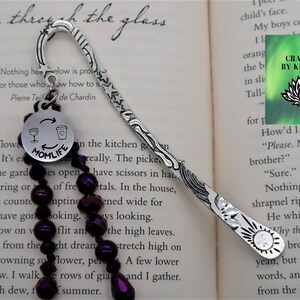 May include: Silver bookmark with a decorative design and a purple beaded chain. The bookmark has a charm with the words "Momlife" and a wine glass and coffee cup graphic. The text "Crafted by Kristal" is visible on a green background with a lotus flower graphic.