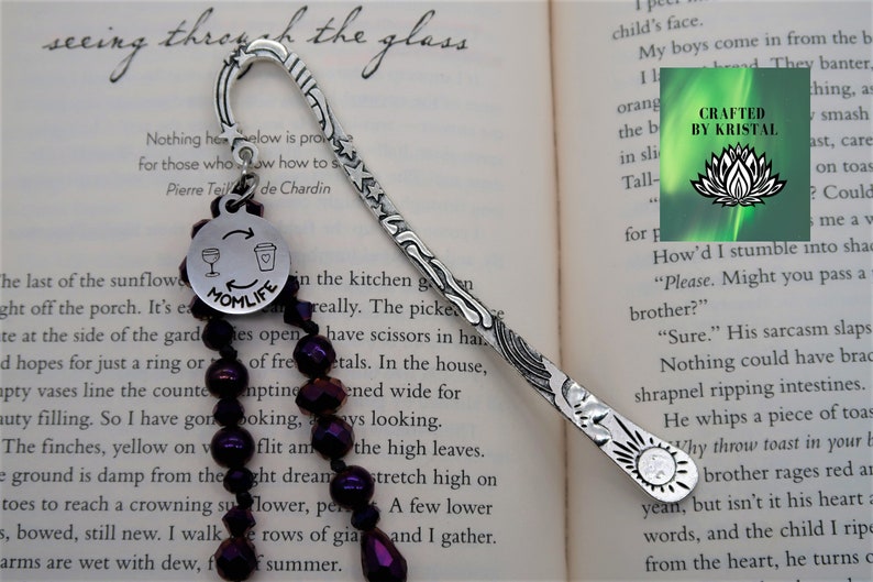 May include: Silver bookmark with a decorative design and a purple beaded chain. The bookmark has a charm with the words "Momlife" and a wine glass and coffee cup graphic. The text "Crafted by Kristal" is visible on a green background with a lotus flower graphic.