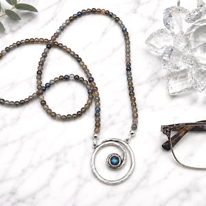 May include: A long beaded necklace with a silver pendant featuring a spiral design and a blue gemstone. The necklace is composed of small, round, gray and blue beads. A pair of eyeglasses and a decorative crystal are also visible.