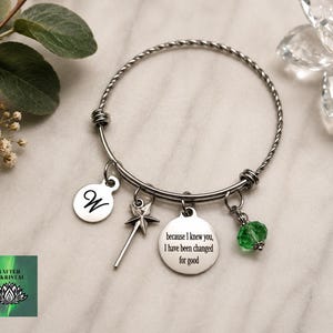 May include: Silver-toned bangle bracelet with multiple charms. Charms include a disc with the letter "W", a star-shaped wand, a disc with the text "because I knew you, I have been changed for good", and a green crystal bead. The bracelet is on a marble surface.