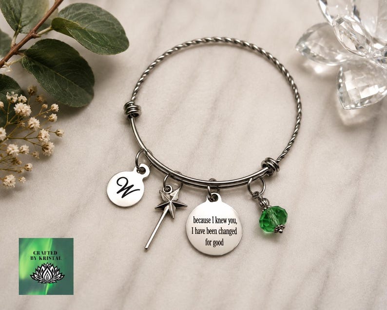 May include: Silver-toned bangle bracelet with multiple charms. Charms include a disc with the letter "W", a star-shaped wand, a disc with the text "because I knew you, I have been changed for good", and a green crystal bead. The bracelet is on a marble surface.