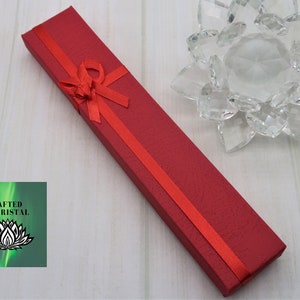 May include: A red gift box with a red ribbon and bow. The box is made of cardboard and has a textured surface. The box is sitting on a white wooden surface. The text "CRAFTED BY KRISTAL" is visible on a green background.