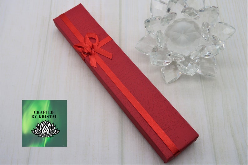 May include: A red gift box with a red ribbon and bow. The box is made of cardboard and has a textured surface. The box is sitting on a white wooden surface. The text "CRAFTED BY KRISTAL" is visible on a green background.