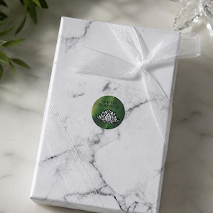 May include: A rectangular gift box with a white marble pattern, tied with a sheer white ribbon. A green circular sticker with the text "Crafted by Kristal" and a lotus flower design is affixed to the box. The box is on a white surface.