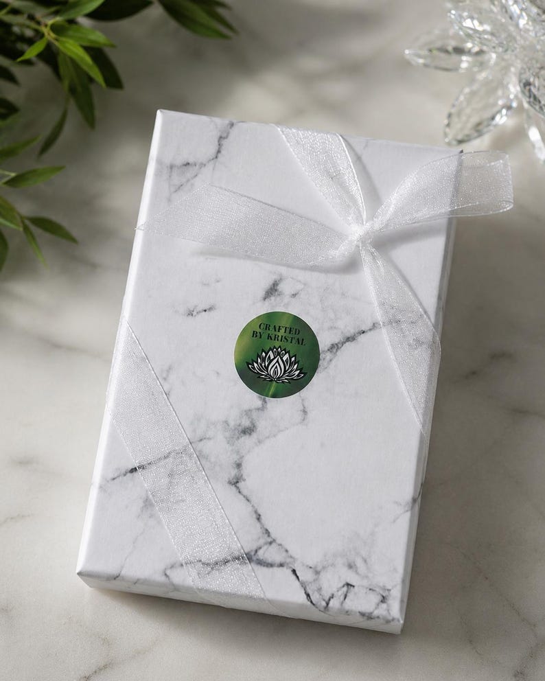 May include: A rectangular gift box with a white marble pattern, tied with a sheer white ribbon. A green circular sticker with the text "Crafted by Kristal" and a lotus flower design is affixed to the box. The box is on a white surface.