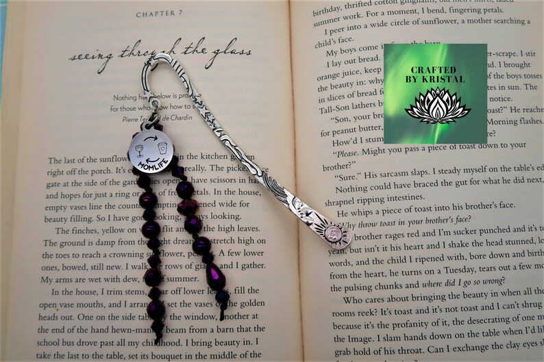 May include: Silver bookmark with a charm that says "Momlife" and a glass of wine with a wine bottle. The charm is attached to a strand of dark purple beads. The bookmark is laying on a book with the text "seeing through the glass" and "Chapter 7".