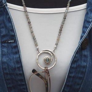 May include: A silver-toned necklace with a spiral pendant holding eyeglasses. The necklace features a beaded chain with small, gray-green stones. The pendant has a small, round, blue-green stone in the center. The necklace is worn over a white shirt and a denim jacket.