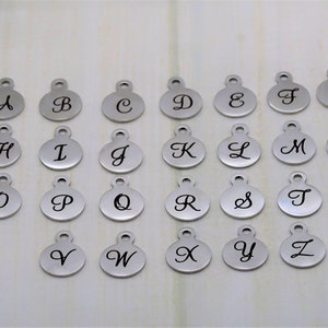 May include: A collection of silver-colored alphabet charms, each with a cursive letter from A to Z. The charms are round with a small loop at the top for attaching to jewelry or other crafts. The letters are in black cursive.