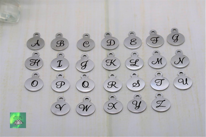 May include: A collection of silver-colored alphabet charms, each with a cursive letter from A to Z. The charms are round with a small loop at the top for attaching to jewelry or other crafts. The letters are in black cursive.