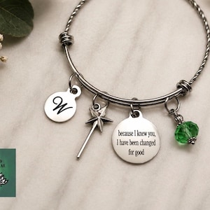May include: A silver-toned bangle bracelet with multiple charms. Charms include a disc with the letter "W", a star-shaped wand, a disc with the quote "because I knew you, I have been changed for good", and a green crystal bead. The bracelet is on a marble surface.