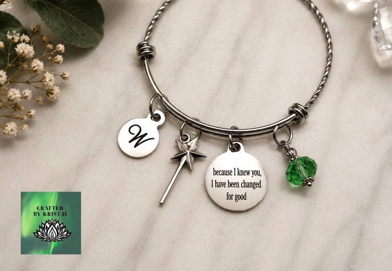 May include: A silver-toned bangle bracelet with multiple charms. Charms include a disc with the letter "W", a star-shaped wand, a disc with the quote "because I knew you, I have been changed for good", and a green crystal bead. The bracelet is on a marble surface.